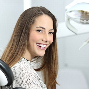 A young, teenage female with long, dark hair smiles while sitting in the dentist’s chair after receiving dental sealants
