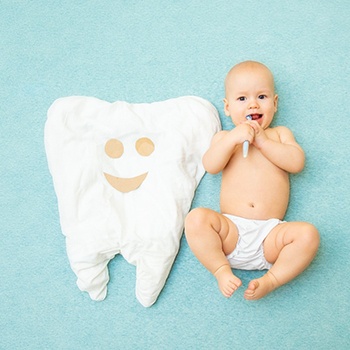 A baby holding a toothbrush lying next to a large tooth pillow