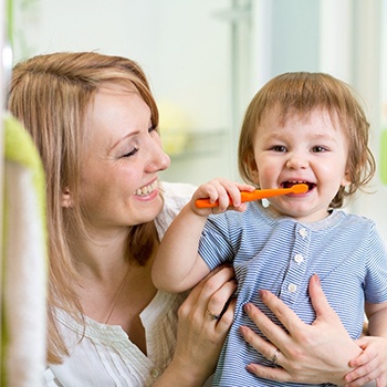 A young toddler brushing their teeth while their mother holds them close and smiles